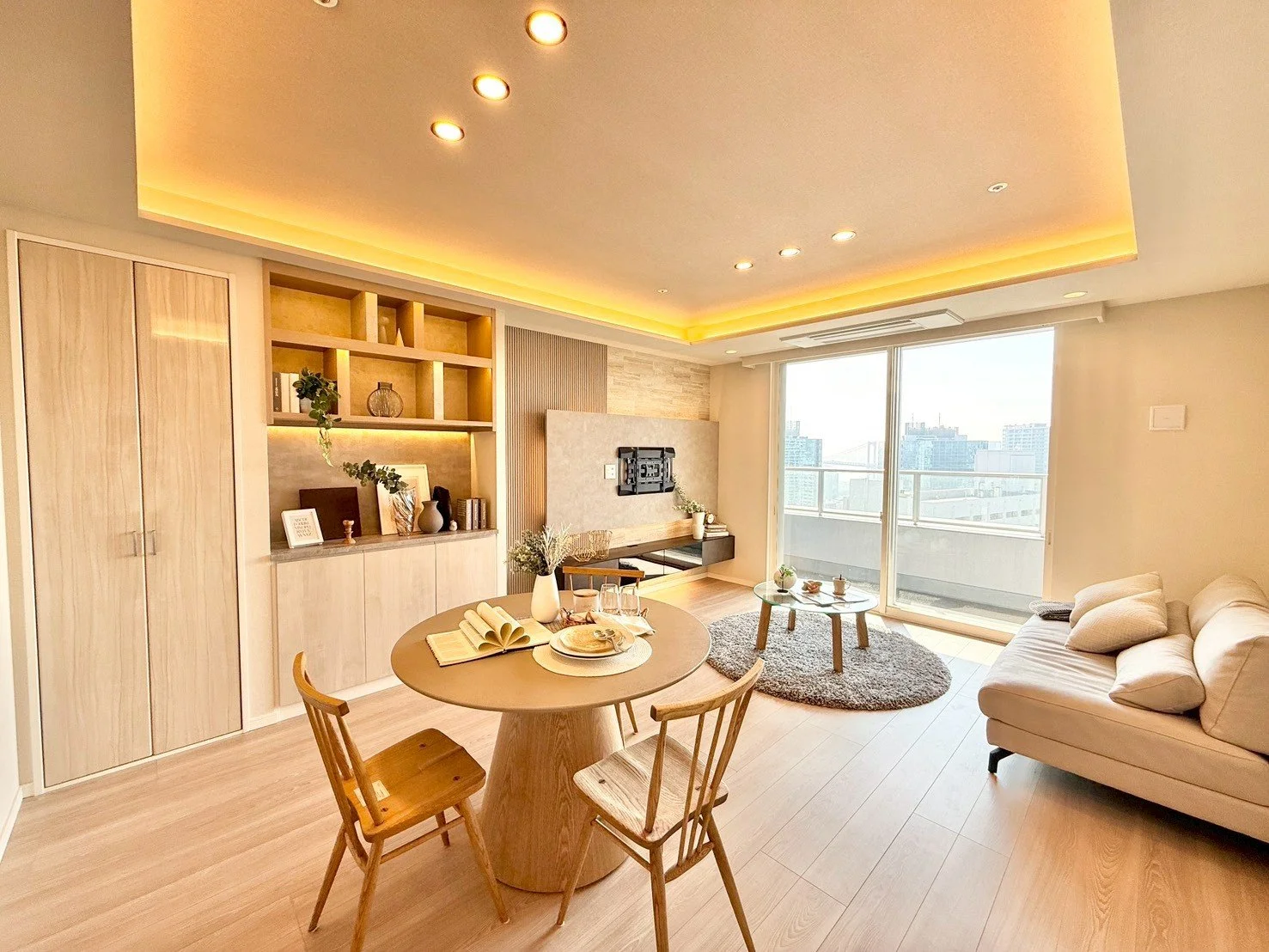 明るいリビングの室内、ソファ、ローテーブル、窓の外に都市の景色が見える、暖色の間接照明が天井に設置されている。