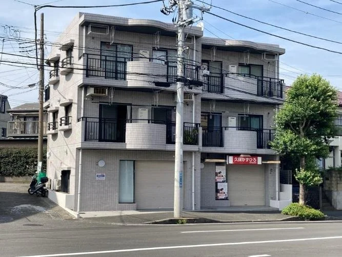 三階建てのコンクリート建築のアパートメント。1階にはシャッターがおりている店舗。道路と電柱が前にあり、左側にバイクが停まっている。右側には木の木々が生えている。