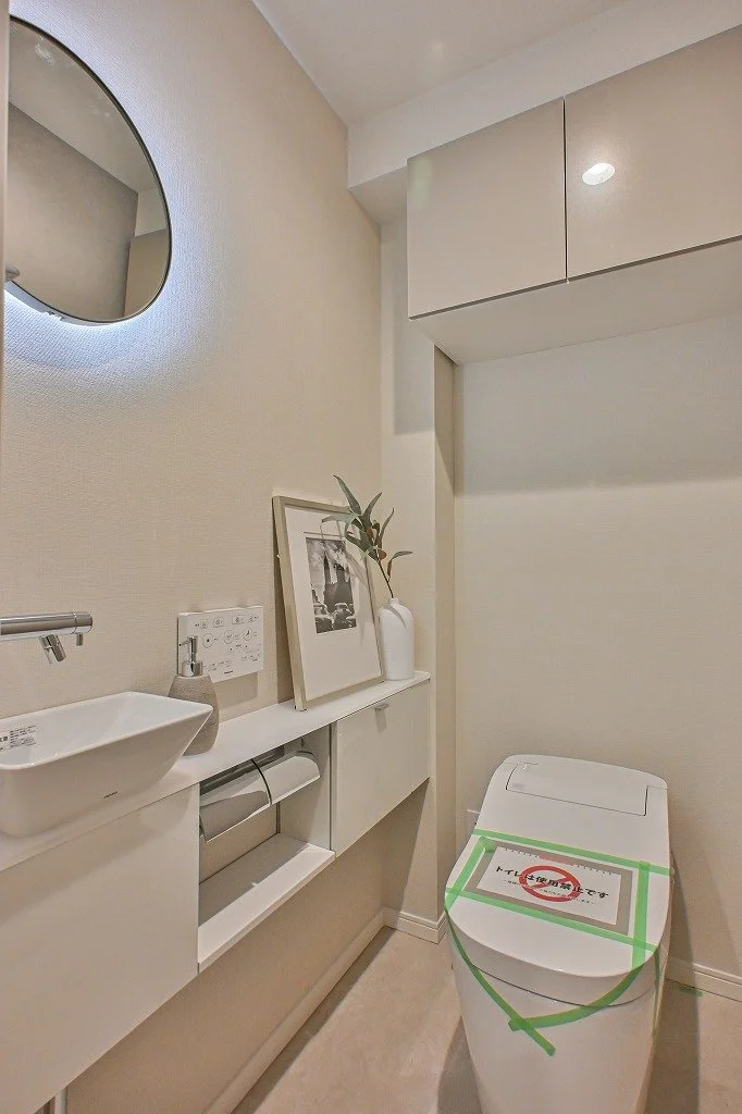 白い壁と家具が配置されたトイレの室内。左側に丸い鏡と小さな洗面台、その下にトイレットペーパーホルダーと紙が置かれている。中央にはブラック＆ホワイトの写真の飾りと植物入りの白い花瓶が配置されている。右奥には蓋つきの便器があり、緑のテープと紙で封鎖されている。