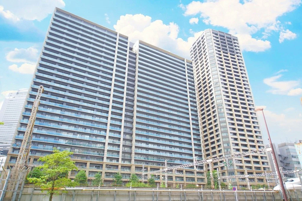 高層の都市型マンションとビルが見える都市の景色、青空と雲が背景にあり、一部に緑の木も見える。