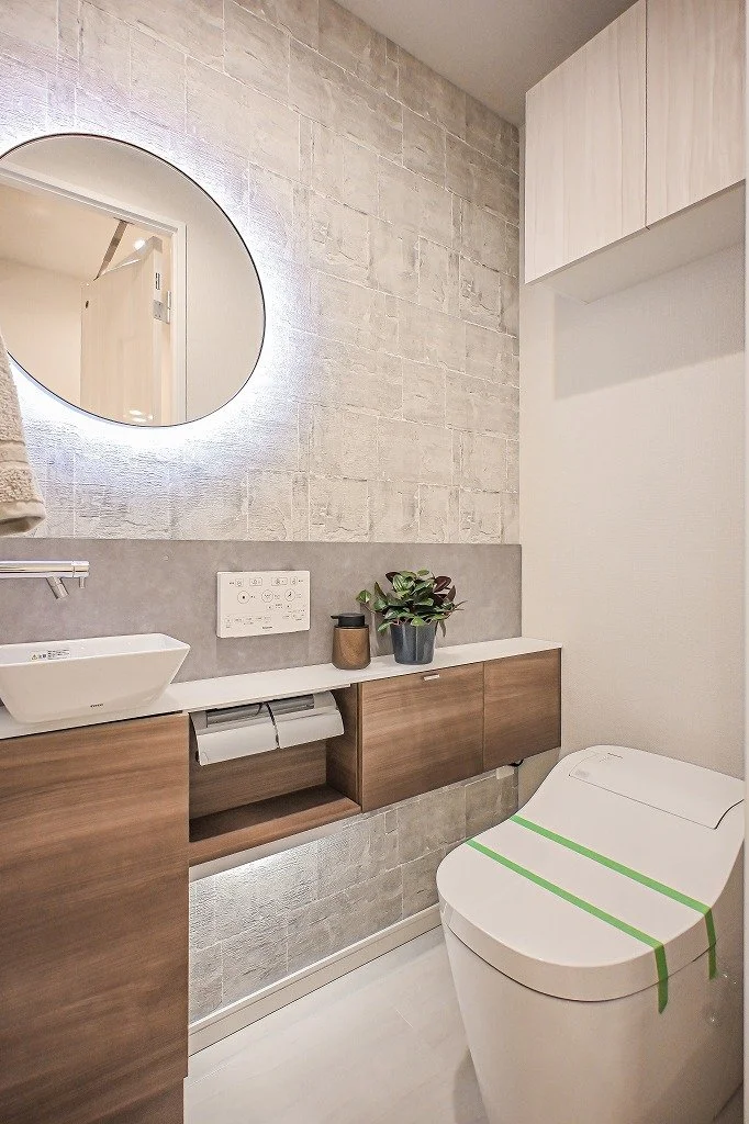 現代的なトイレルームで、丸い鏡と洗面台、植物と収納棚がある。