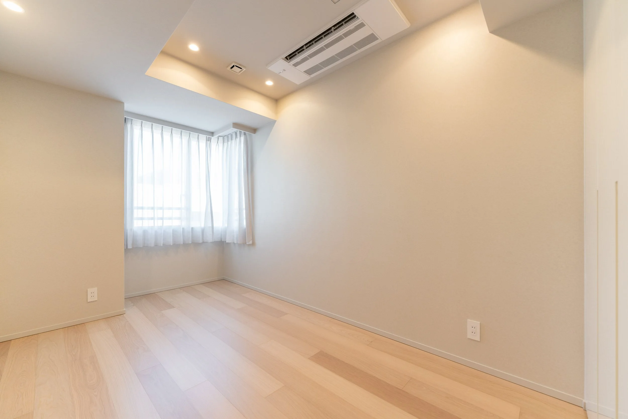 白い壁と木のフローリングが特徴の空間。窓には白いカーテンがかかっており、天井にはエアコンと照明が設置されている。