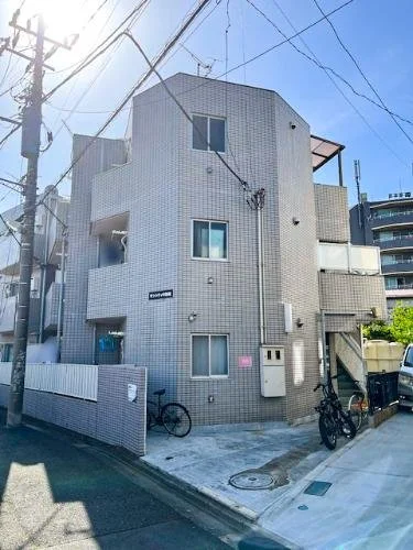 三階建ての白いレンガのアパートメント。自転車と電気ボックスが前にあり、電柱と電線も写っている。