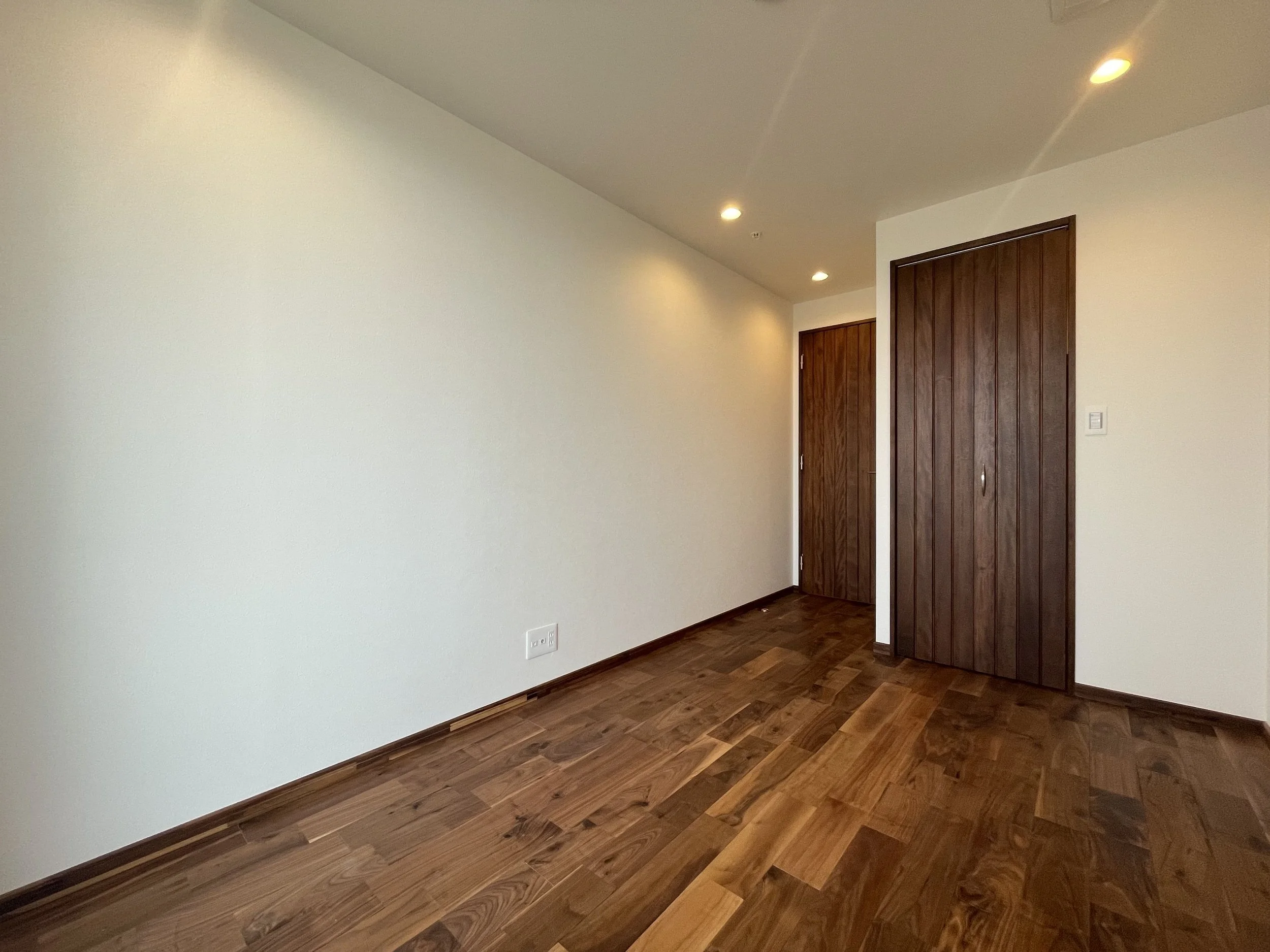 木製の扉が二つある空室の部屋。白い壁と木の床。照明の天井灯が点いている。
