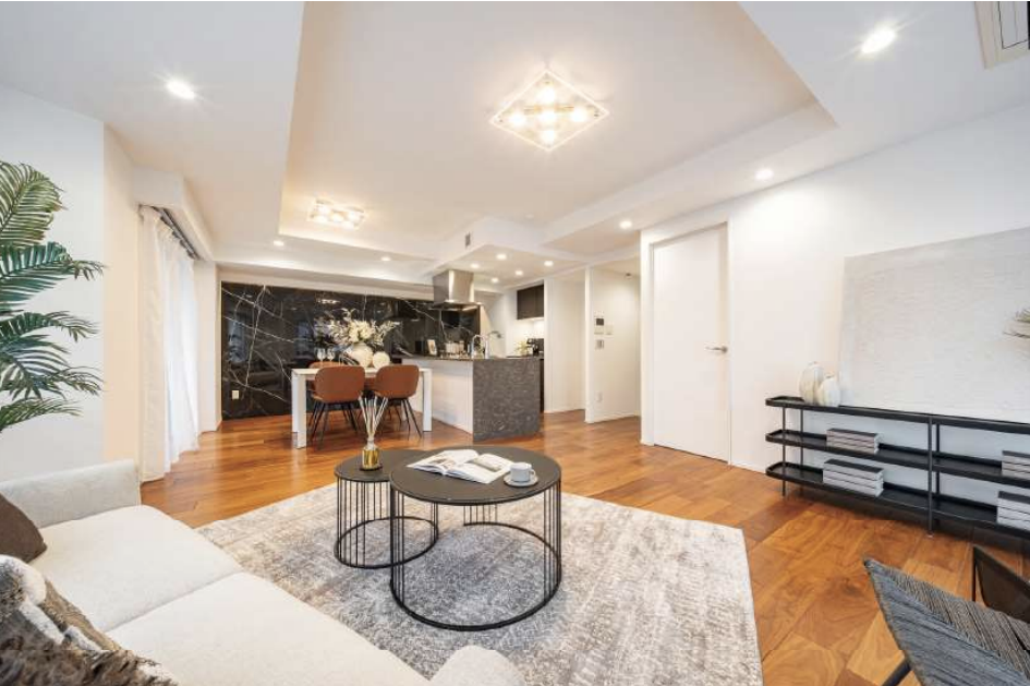 リビングルームとダイニングエリア、キッチンが見える現代的な居住空間の写真。木製フローリングと白い壁が特徴で、ソファやテーブル、棚などの家具が配置されている。