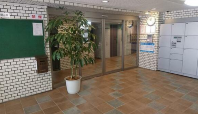ロビーのようなエリア、植物の鉢、掲示板、郵便ポスト、受付またはロッカー、エレベーターの前のガラス扉、壁に時計と掲示板がある。