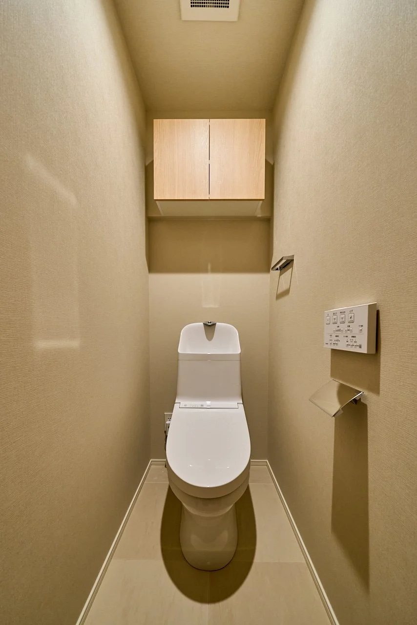 狭いトイレの中にある洋式便器と便座横の操作パネル、背面の木製キャビネット。