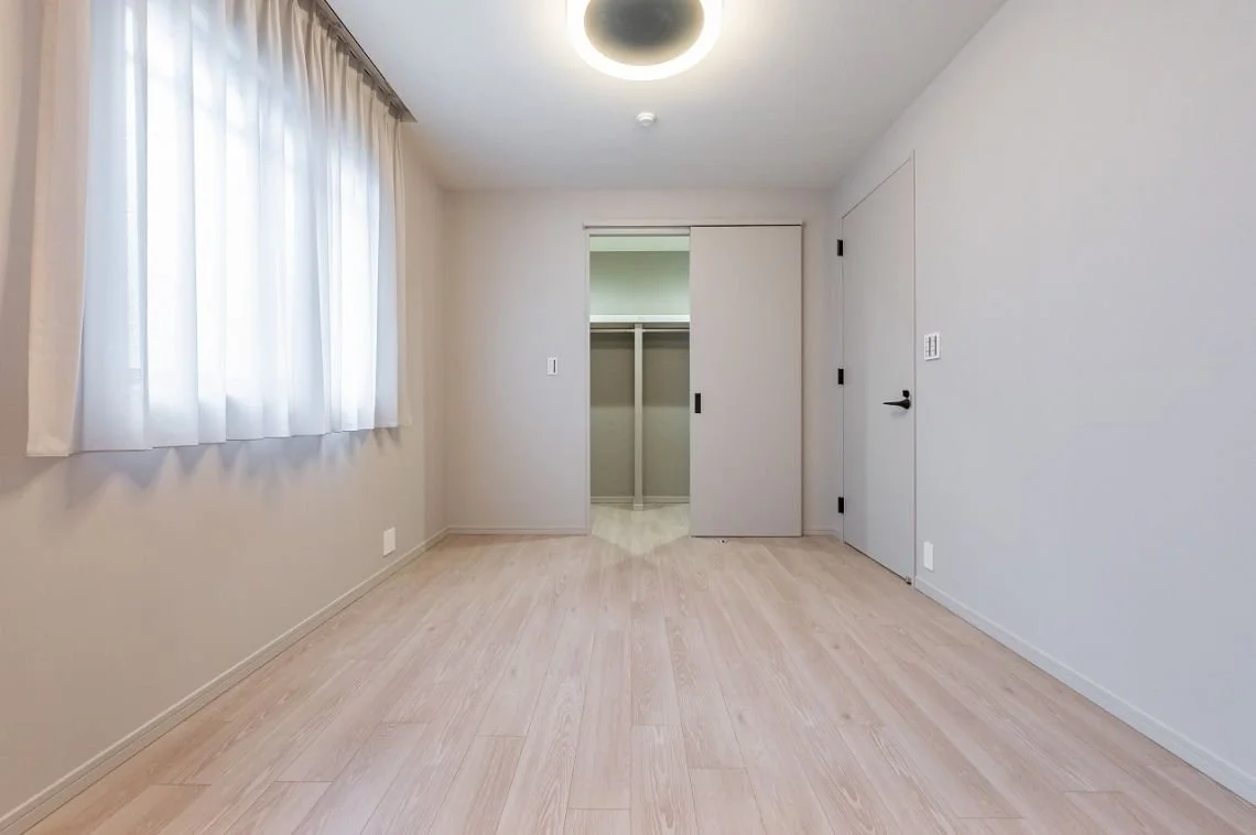 白い壁と木製フローリングの空室、窓に白いカーテンが掛かっている。左右には扉があり、中にはクローゼットが見える。