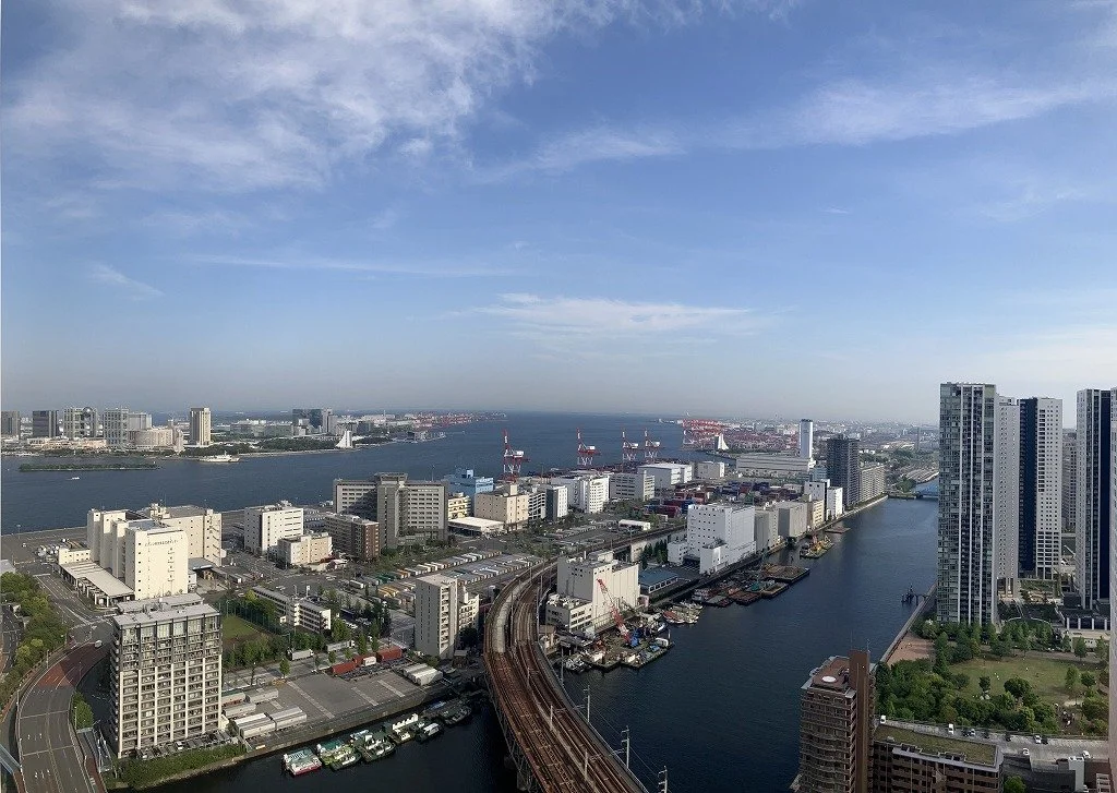 都会の川沿いに高層ビルと工場が立ち並ぶ都市景観。青い空と白い雲も見える。」}
