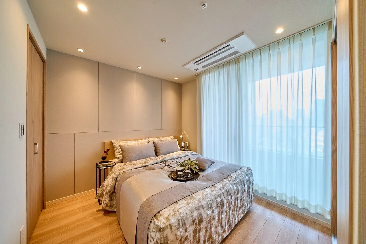 明るい窓際のベッドルーム、木目調の床とシンプルな壁紙、窓には白いカーテンが掛かっている。ベッドに枕と毛布、トレイに植物とカップが置かれている。