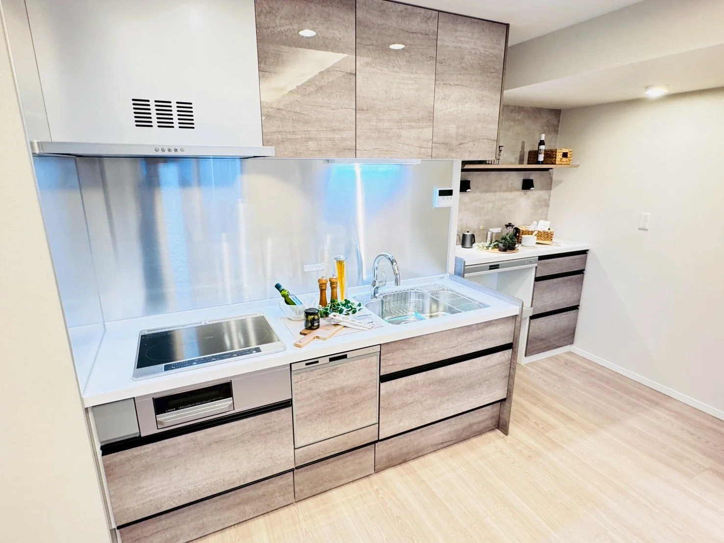 モダンなキッチンで、白いカウンターと木目調のキャビネット、ステンレス製のバックパネルとレンジフードが特徴。中央にはシンクと調理器具が配置され、背面には棚や調味料、装飾品が置かれている。