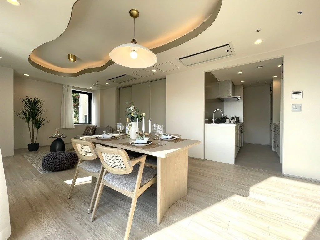 明るくモダンなリビングとダイニングエリア。木製の家具、窓からの日光、植物、シンプルな装飾が施されている。キッチンには白いキャビネットとステンレスの設備が見える。