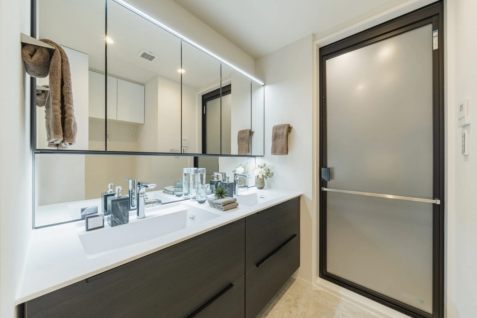 白い洗面台と上部に鏡、タオル、花の飾りが置かれた洗面所の内装。収納キャビネットとスライドドアも見える。