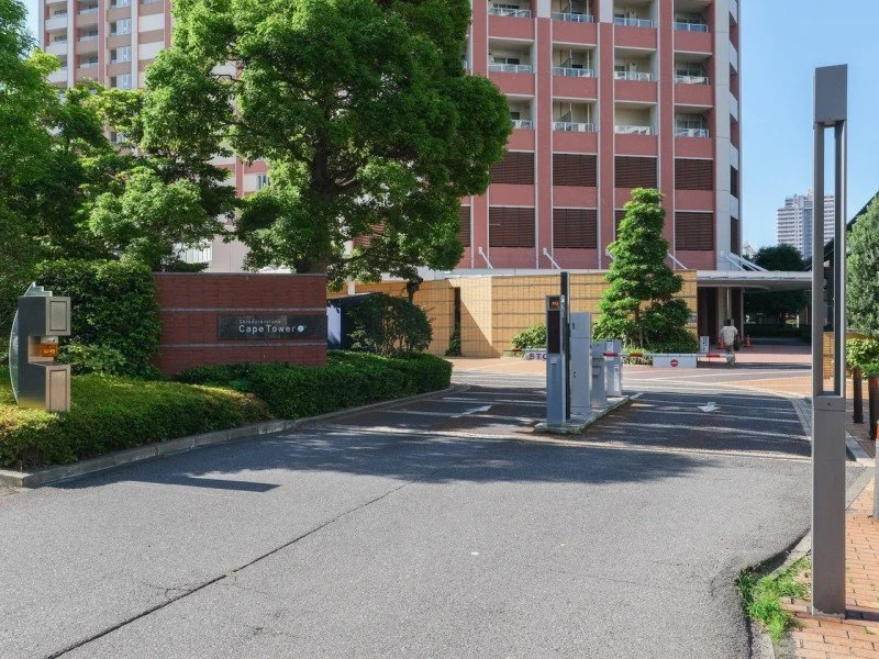 高層マンションの入口にあるゲートと駐車場、自動車の出入り口が見える。左側には緑の木と植栽、建物の名前の看板が見える。