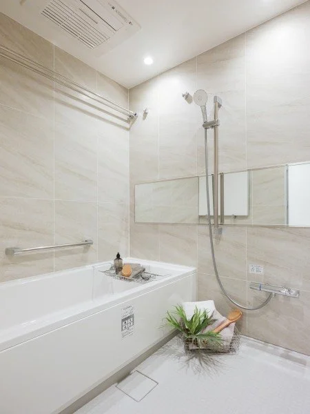 白い浴槽とシャワー付きのバスルームの壁に鏡が設置されている。バスルームには緑の植物とバス用品が置かれている。