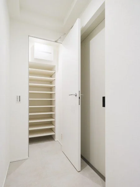 白い棚が並ぶ狭い収納スペースとドア、エレベーターの扉のある室内