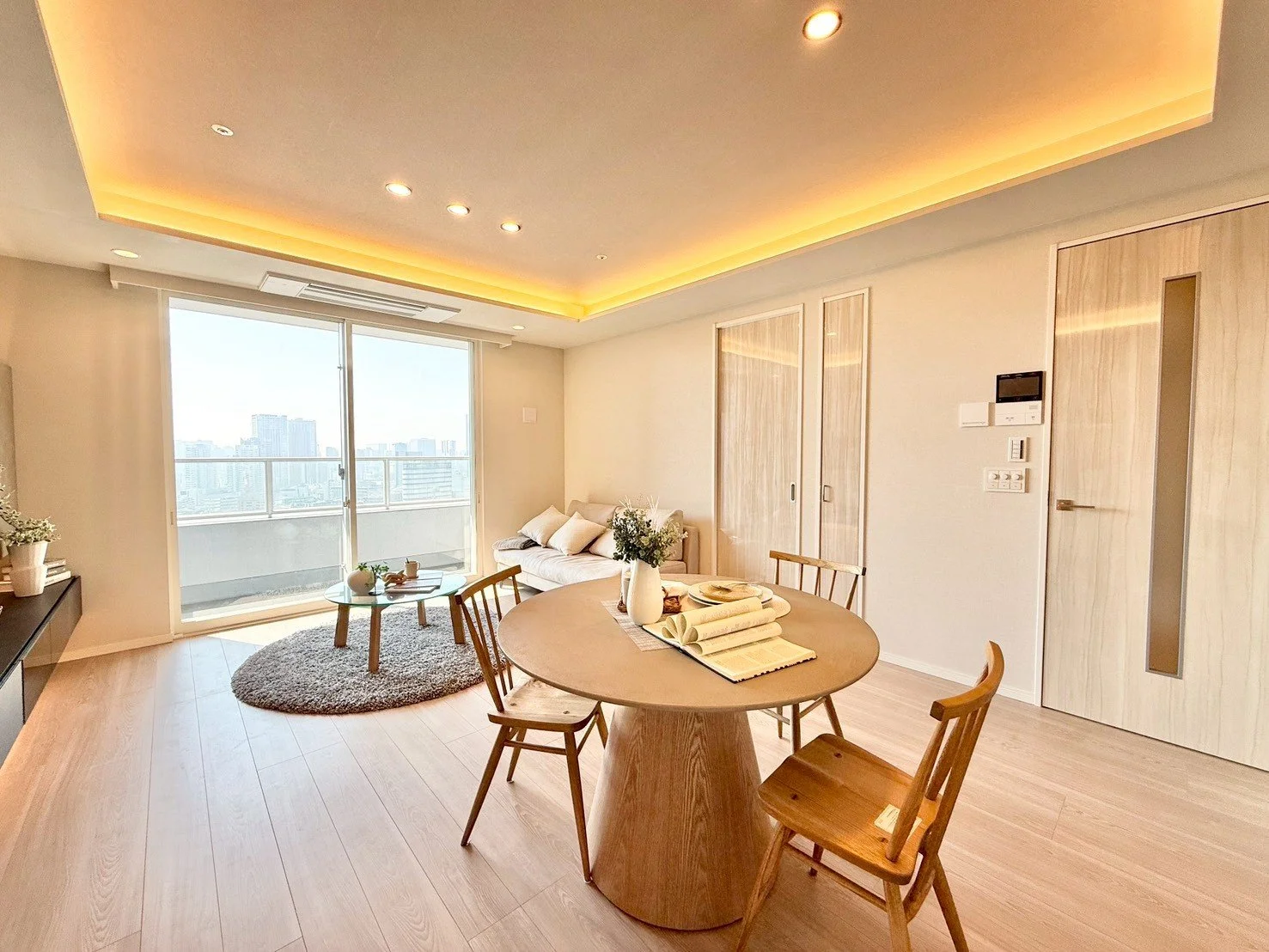 明るく開放的なリビングルームとダイニングエリア、ソファと丸いテーブル、バルコニーに面した大きな窓から眺める都会の景色。