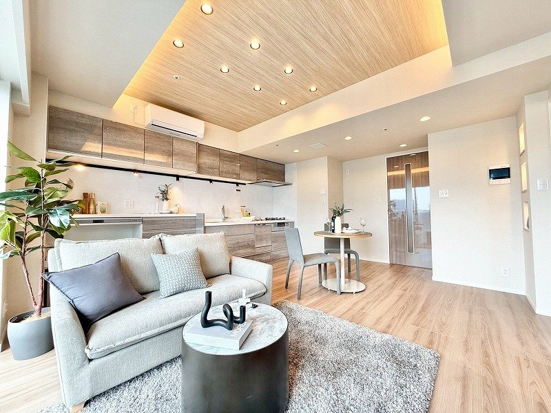 モダンなリビングルームとキッチンのオープンスペース、ソファーとダイニングテーブル、木目調の天井とカウンタースペース、観葉植物あり。