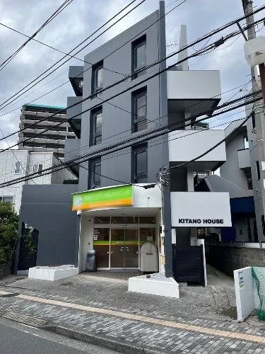 三階建ての黒い建物で、下はコンビニのKITANO HOUSE。建物の前に電柱と電線があり、道路にはコンクリートブロックの付いた歩道がある。