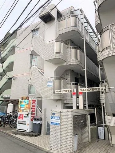 都市のアパートメントビルの外観。自転車と自動販売機が前にあり、看板やバルコニーが見える。