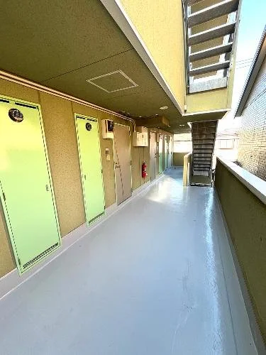 建物の階段付きの廊下の眺め。左側には緑色のドアが並び、右側に外壁の柵と階段が見える。