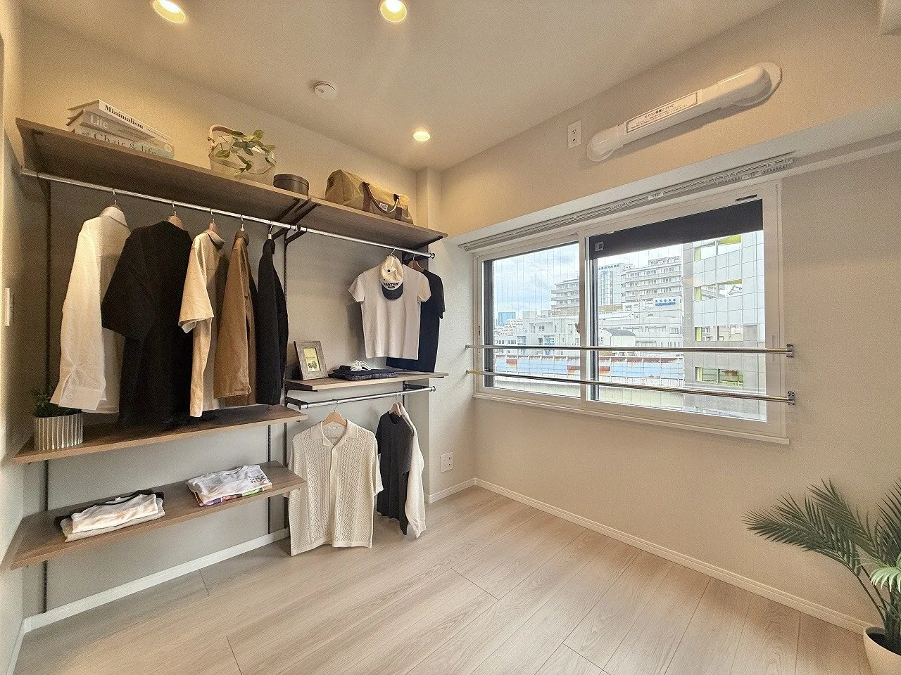 シンプルな部屋。壁にハンガーラックと棚が設置され、衣服と雑貨が掛かっている。右側の大きな窓から都市の景色が見える。室内には植物も置かれている。