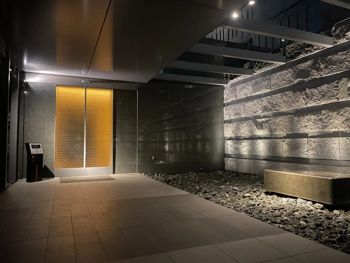 夜のエントランス、ガラスドア、壁に照明の陰影、岩とタイルの装飾、ベンチとインターホン