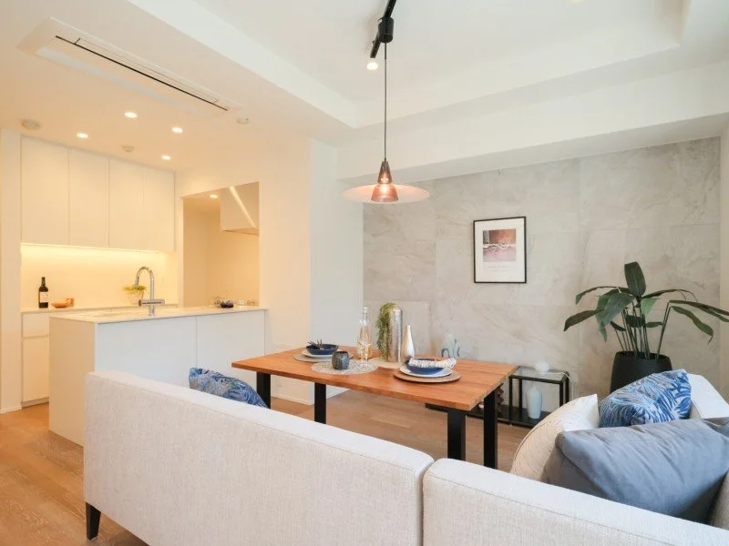 白いリビングルームのダイニングエリアと小さなキッチン。木製テーブルに皿やカップ、ガラス製品が置かれ、後ろにはソファと観葉植物が見える。シンプルで明るいインテリア。