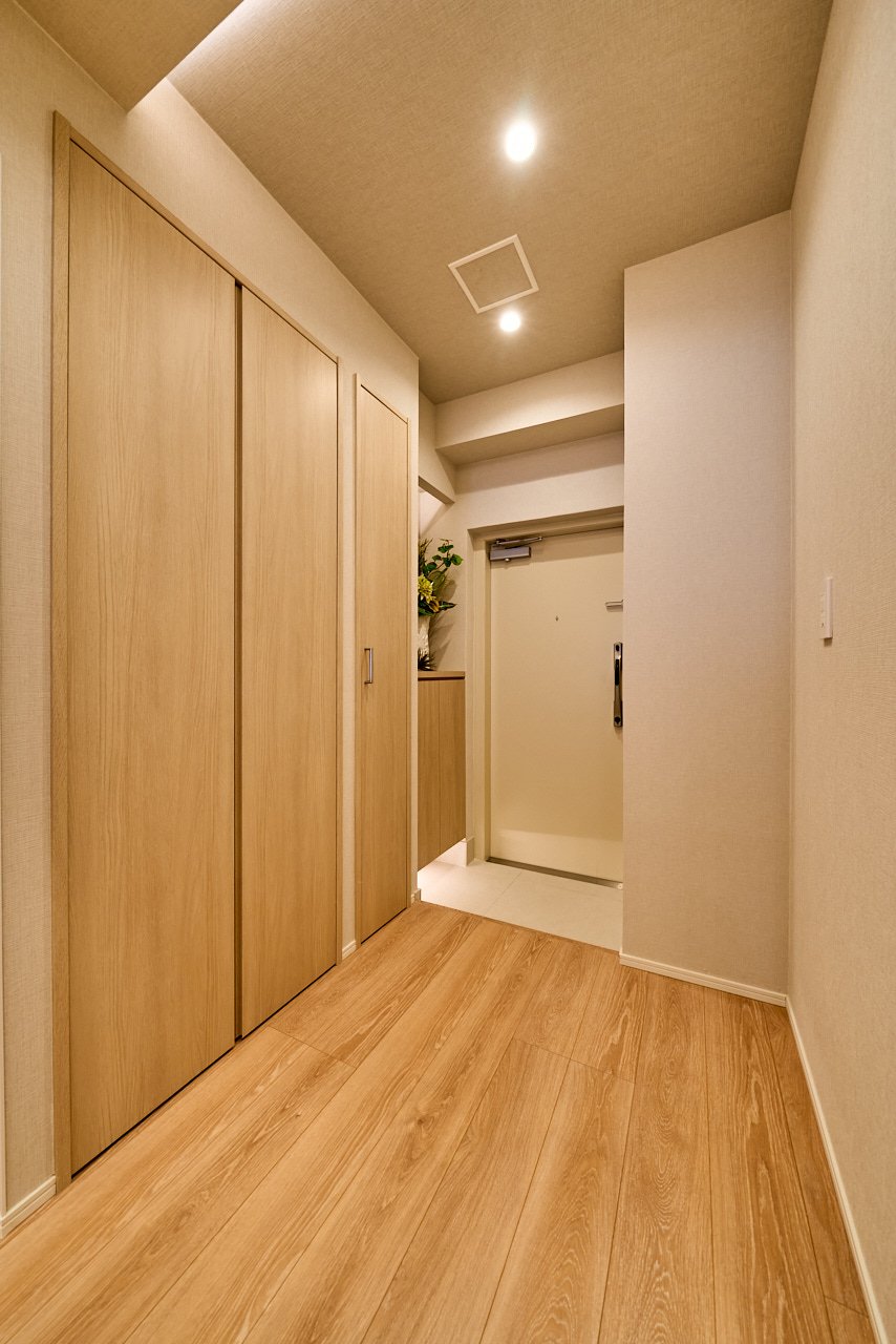 木製の扉が並ぶ玄関のホール。扉の横には花の飾りや収納棚があり、天井に照明と換気扇が見える。