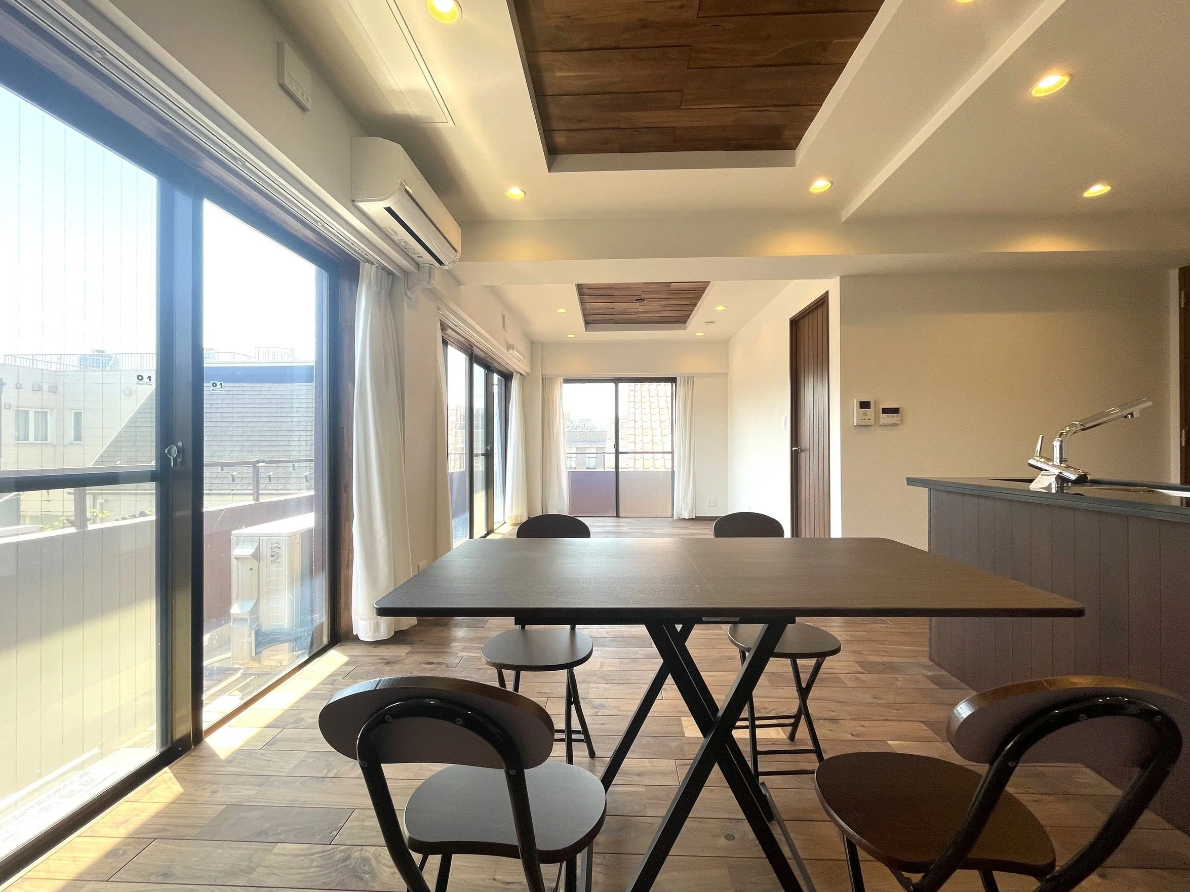 明るいリビングルームの interior with large glass doors,ダイニングテーブルと椅子があり、木製の床と天井の一部に木材のデザインが施されている。自然光が差し込み、都市の景色が見える。