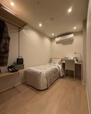 シンプルなベッドルームの部屋で、ベッド、デスクと椅子、エアコンが設置されている。