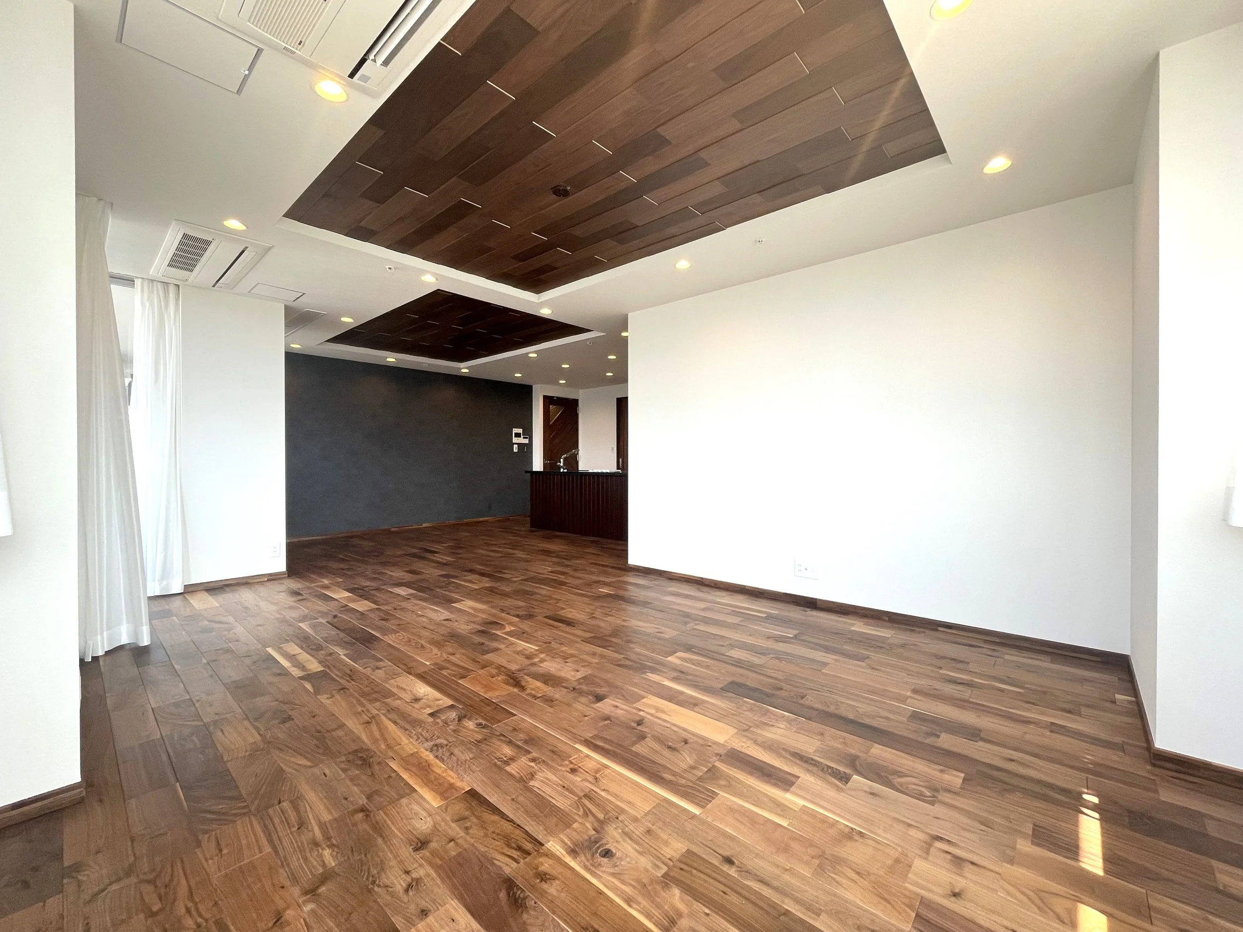 木製の床と天井のデザインが特徴的な広い空間、白い壁とダークなアクセントウォール、窓に白いカーテンが掛かっている