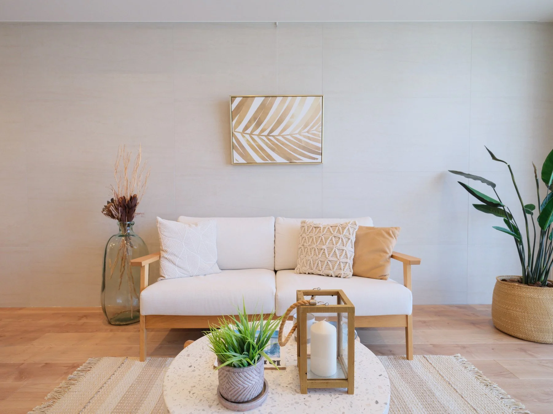 シンプルでナチュラルなリビングルーム。白いソファと木製のコーヒーテーブル、クッション、観葉植物や装飾が配されている。壁には葉のアートが飾られ、温かみのあるウッドフロア.