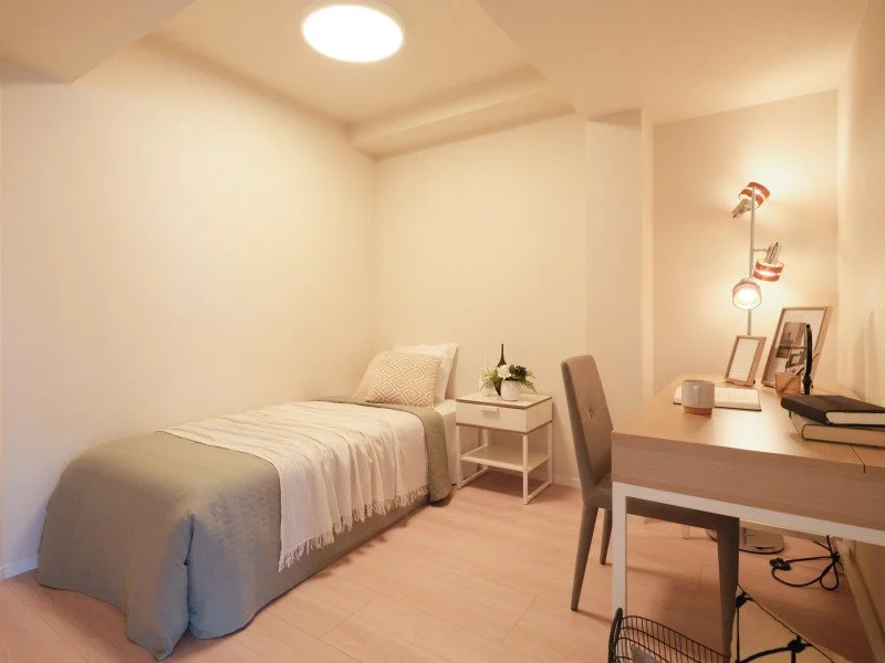 白い壁の部屋で、シングルベッドとデスク、椅子、ランプがあります。