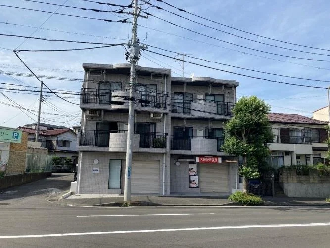 三階建てのアパートメントの建物。道路の前に電柱と電線があり、いくつかのバルコニー付きの部屋が見える。右側に木がある。
