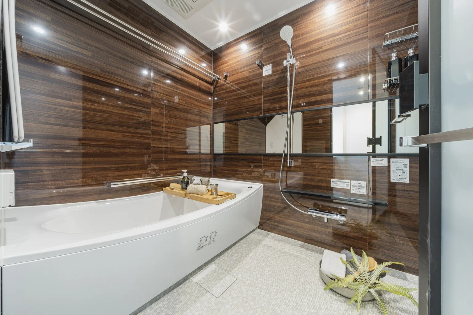 木目調の浴室内に白い浴槽と鏡、シャワーヘッド、サウナ用の椅子や植物が配置されたモダンな浴室