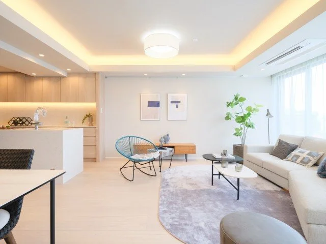 明るいリビングルームとダイニングエリアの室内風景。白いソファとコーヒーテーブル、青い椅子、観葉植物があり、柔らかい照明が天井から照らしている。