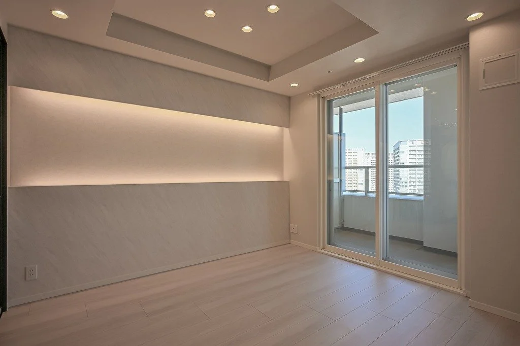 明るいリビングルーム、白い壁と木製の床、天井には埋め込み照明、ガラスのバルコニー扉と外の都市景観が見える