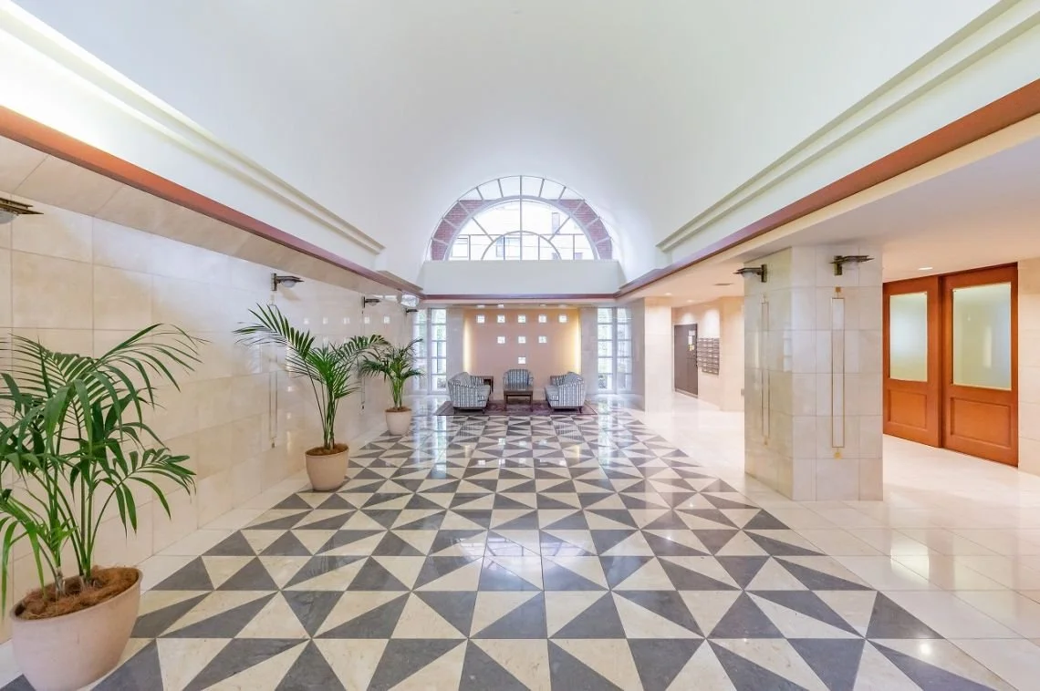 建物のエントランスホール、白と黒の幾何学模様のタイル床、観葉植物の鉢、向こう側に三脚の椅子とテーブル、丸い窓とガラスブロックの窓