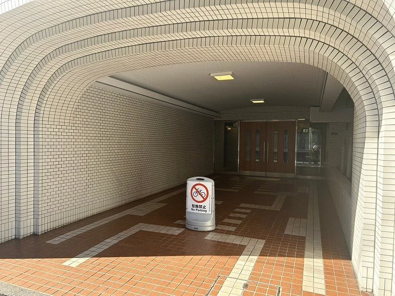自動販売機の近くに『駐輪禁止』の看板が立てられた通路の入り口。通路はレンガ風のタイル張りで、壁と天井は白いタイルで覆われている。扉は木製で、窓がついている。