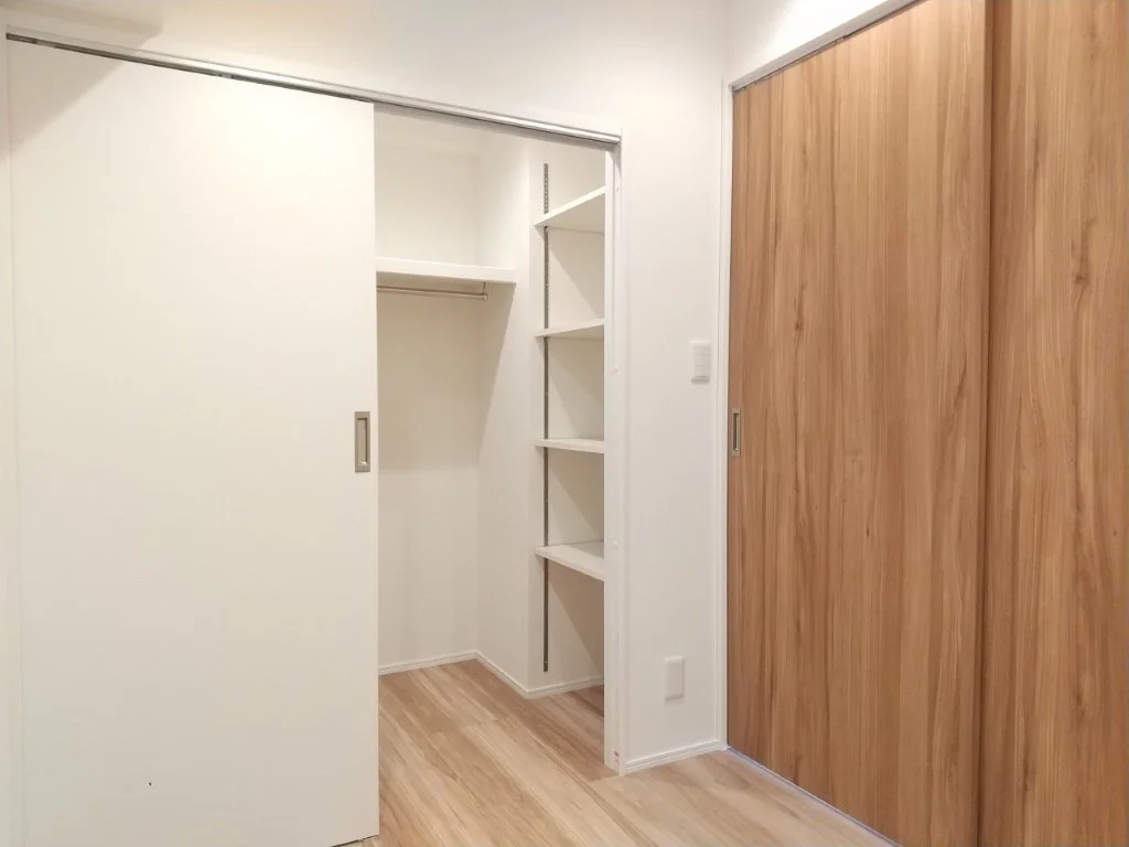 白いクローゼットの中に棚とハンガーパイプがあり、木製の扉のクローゼットも壁側に設置されている室内の収納スペースの写真