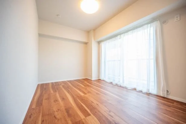 日当たりの良い空の部屋で、木製の床と白い壁、窓にカーテンが掛かっている
