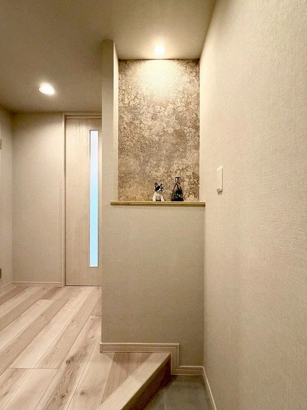 廊下の角に小さな飾り棚があり、ウサギの置物とガラスの瓶が置かれている。背後の壁は模様のあるアクセントウォール。照明が天井に設置されている。