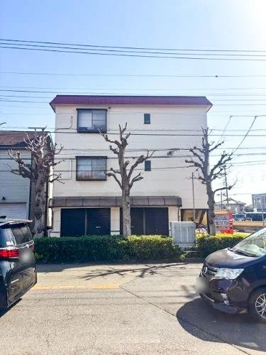 3階建ての白い建物と、前の道路に駐車された車、枝を落とした木々が並ぶ風景