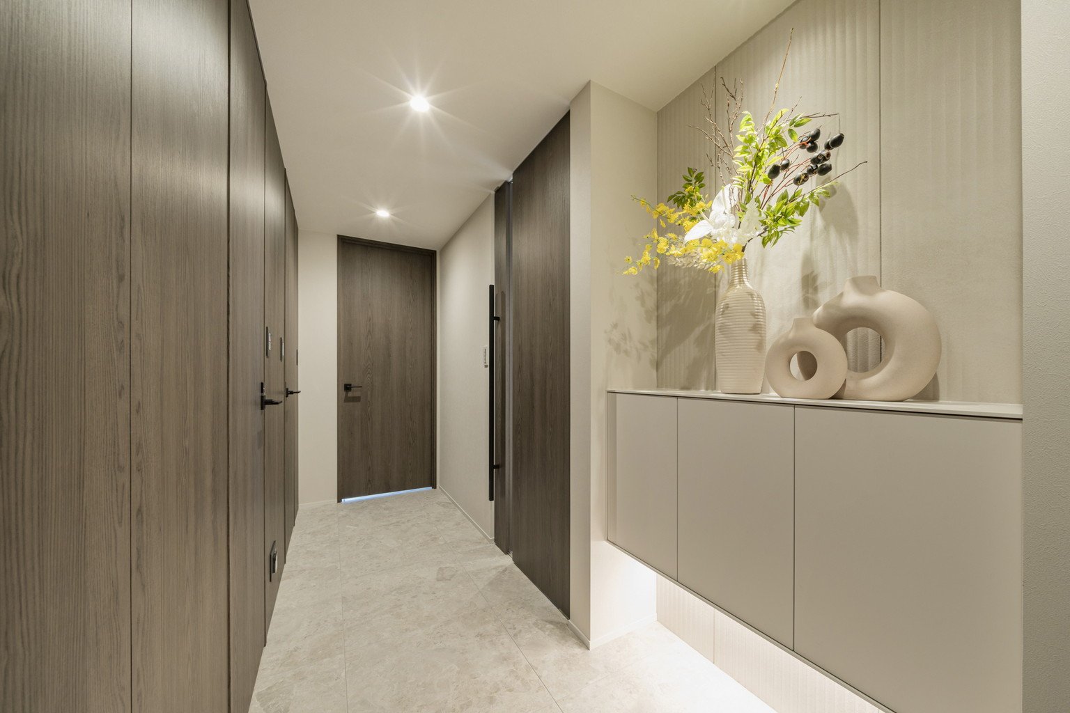 木製の扉とキャビネットがあるモダンな玄関ホール。白い花と植物のアレンジメントが飾られたセラミック製の花瓶と装飾的な白い陶器のオブジェが置かれている。照明が天井に設置されている。