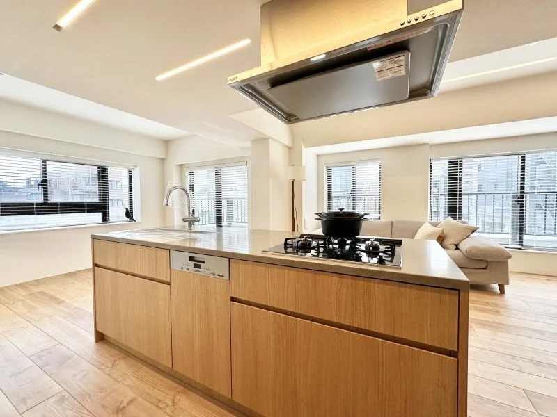 木製のキッチンカウンターとコンロ、レンジフード、ソファが置かれたリビングルーム、複数の窓から自然光が入っている。