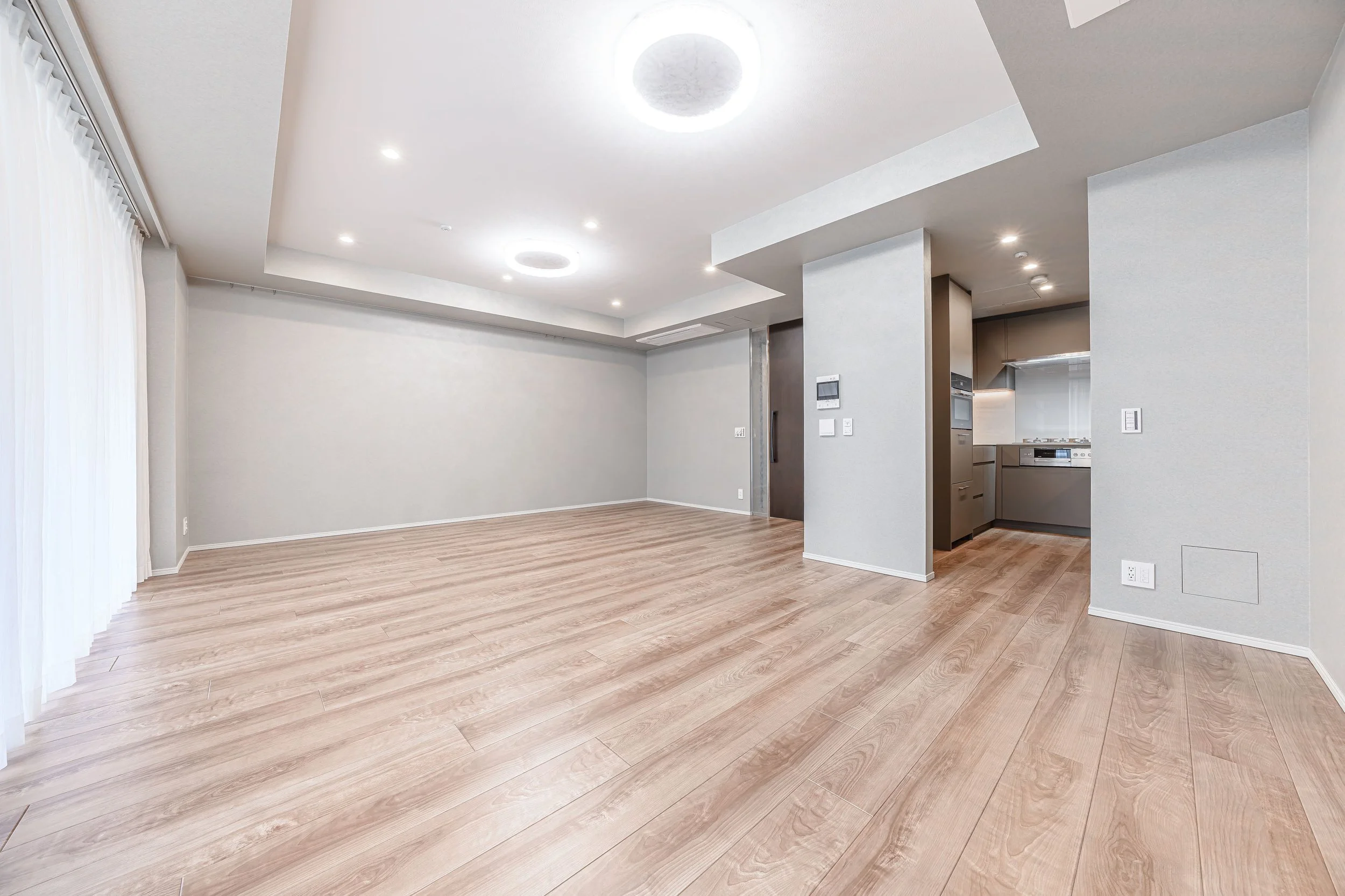 白い壁と天井、木製床の空き部屋、キッチン部分とバルコニー側のカーテンが見える