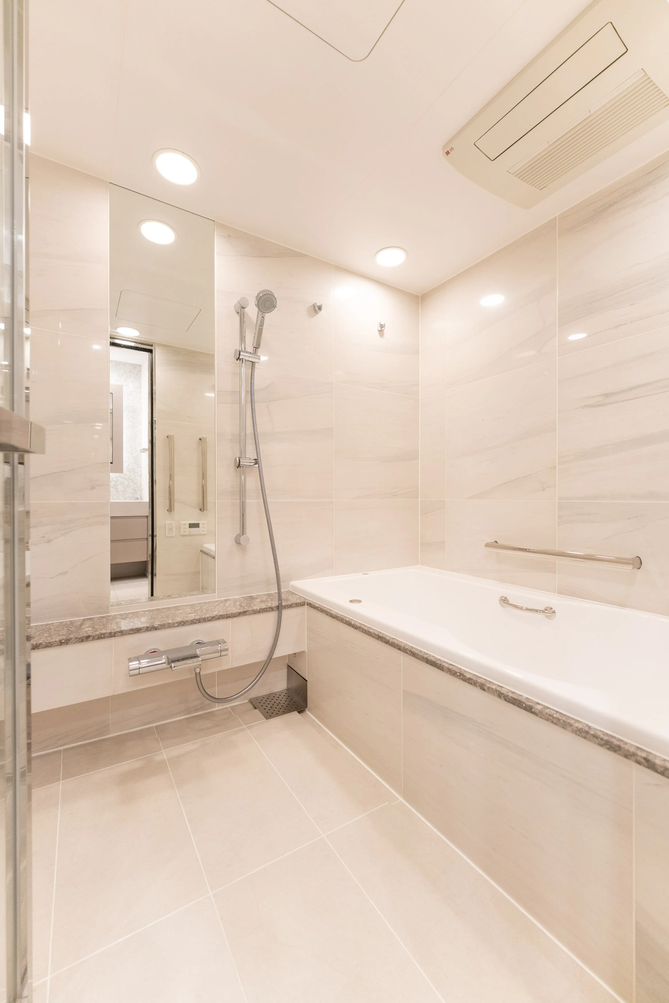 白いタイル張りの浴室。浴槽、シャワー、ミラー、照明が備えられ、清潔な空間。