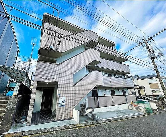 三階建てのアパートメントビル、階段の外側に通路があり、バイクと自転車が駐車されている。空に雲が広がる風景。