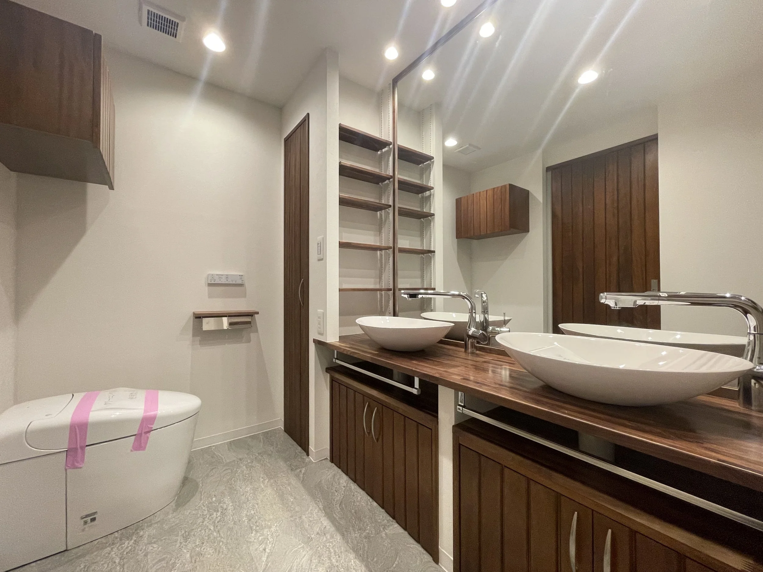 木製の洗面台に2つの白いボウル型洗面器、横に大きな鏡、左側にトイレと木の扉、壁に棚と換気扇、天井にスポットライト照明.